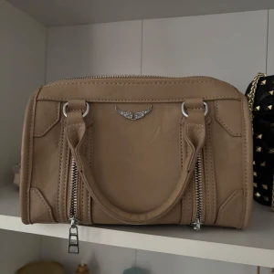 Beige handväska med silverdetaljer - Snygg beige handväska i skinn med två dragkedjor framtill och ett emblem med glittrande stenar. Väskan har rundade former och dubbla handtag, zadig liknande väska