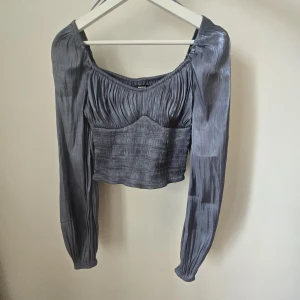 Grå glansig blus med smock och puffärm - Supersnygg grå blus från Bik Bok i glansigt material med smockad midja och puffiga långa ärmar. Toppen har rynkade detaljer upptill och en croppad passform som ger en trendig look. Perfekt för dig som vill sticka ut lite extra.