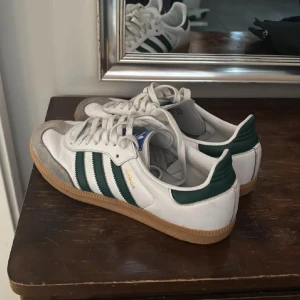 Adidas Samba vita och gröna sneakers - Klassiska Adidas Samba sneakers i vitt skinn med gröna detaljer och de ikoniska tre ränderna på sidan. Skorna har gummisula, snörning och rund tå. Perfekta för dig som gillar retrostil och streetwear.