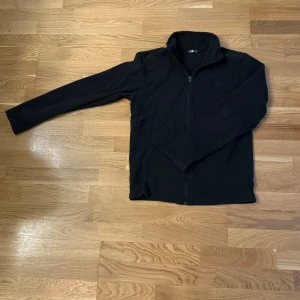 Svart fleecejacka från The North Face - Svart fleecejacka från The North Face med hel dragkedja och hög krage. Perfekt för kyliga dagar, enkel att slänga på över en t-shirt. Klassisk och stilren design med diskret logga på bröstet. Långärmad och riktigt mjuk.