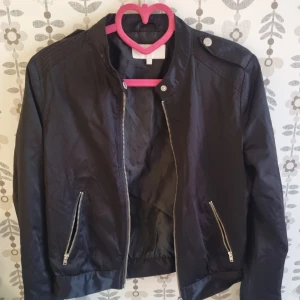 Svart bomberjacka med dragkedjor - Snygg svart bomberjacka med silvriga dragkedjor framtill och på fickorna. Jackan har en klassisk krage med knappdetalj på axeln och raka ärmar. Perfekt för dig som vill ha en stilren och enkel look.