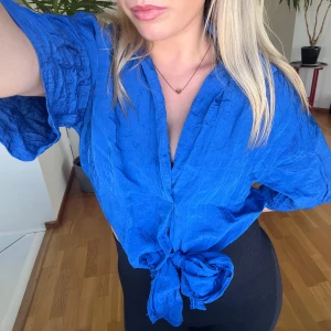 Blå omlottblus med knyt - Säljer en blå omlottblus med korta, vida ärmar och knyt framtill. Blusen har en djup v-ringning och är tillverkad i ett lätt, skrynkligt tyg som ger en avslappnad känsla. Perfekt för dig som vill ha en färgstark och trendig topp.