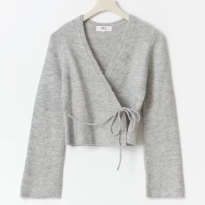 Grå omlottkofta med knyt från Gina Tricot - Supermjuk grå omlottkofta från Gina Tricot med knyt i sidan. Koftan är stickad, har lång ärm och croppad passform. Köpt för 399kr men har aldrig används💕.