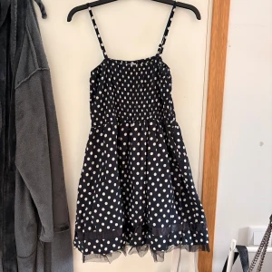 Svart prickig kortklänning Gina Tricot - Supersöt svart kortklänning från Gina Tricot med vita prickar och smockad överdel. Klänningen har tunna axelband, volangkant nertill och ett band i satin vid fållen. Perfekt för dig som gillar lekfulla mönster och lätt material.