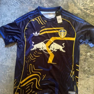 Leeds United x Red Bull Adidas tröja - Snygg fotbollströja från Adidas i samarbete med Leeds United och Red Bull. Tröjan är mörkblå med gula och blå grafiska detaljer, klubbmärke på bröstet och Red Bull-logga framtill. Tillverkad i lätt och ventilerande material, perfekt för match eller träning.
