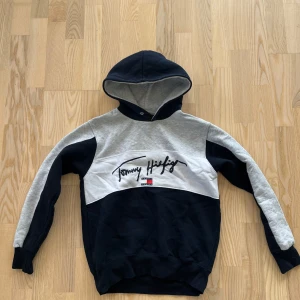 Tommy Hilfiger hoodie i blått och grått - Snygg hoodie från Tommy Hilfiger i mörkblått och ljusgrått med broderad logga framtill. Tröjan har huva, långa ärmar och ribbade muddar. Perfekt för en avslappnad och trendig stil.