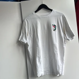 UNIQLO t-shirt med konstverks tryck - Säljer en vit t-shirt med korta ärmar i mjuk bomull. På bröstet finns ett litet tryck av en påfågel och på ryggen ett stort, färgstarkt motiv med blommor och påfågel i rött, grönt och gult. Perfekt för dig som gillar unika prints och vill sticka ut. Ett konstverk av Keiichi Tanaami!