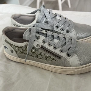 Grå sneakers med blommigt mönster - Snygga grå sneakers från Mustang med blommigt mönster på sidorna, silvriga detaljer och dragkedja. Skorna har snörning, rund tå och platt sula. Materialet är syntet med inslag av skinnimitation. Perfekta för dig som vill ha något unikt och stilrent.