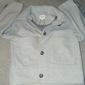 Grå overshirt från Kappahl - Säljer en stilren grå overshirt från Kappahl i storlek S. Jackan har stora bröstfickor, klassisk krage och knäppning med svarta knappar framtill. Materialet är mjukt och känns ulligt, perfekt för lager-på-lager. Enkel och clean look som passar till allt.