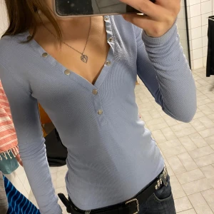 Ljusblå ribbad topp från NA-KD😌😚😚 - Säljer en ljusblå långärmad topp från NA-KD i ribbat material. Toppen har v-ringning med silvriga knappar längs halsringningen och ner mot bröstet. Sitter tight och är perfekt att styla med jeans eller kjol.