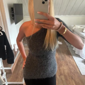 Grå stickad oneshoulder topp - Säljer en supermjuk och fluffig grå stickad topp med oneshoulder-design. Toppen är figurnära och har en snygg asymmetrisk skärning som ger en trendig look. Perfekt att styla med både jeans och kjol för en cool vibe. Prislapp kvar