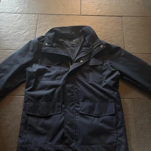 Feldjacket  - Parkajacka i mörkblått från C Studio med fyra stora fickor framtill, dragkedja och tryckknappar. Jackan har hög krage och är tillverkad i ett slitstarkt polyestertyg. Perfekt för kyliga dagar och har en clean, stilren look. Ny pris 1300 