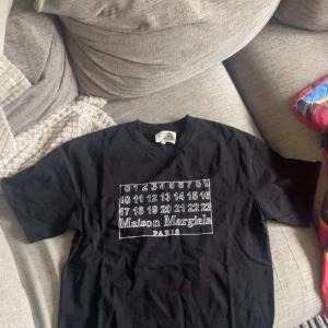 Maison Margiela svart t-shirt - Svart t-shirt från Maison Margiela med ikoniskt tryck framtill med siffror och logga. Klassisk passform, rund hals och korta ärmar. Tillverkad i mjuk bomull för en skön känsla. Perfekt för dig som gillar stilrena och trendiga plagg. Aldrig använd och lappen i nacken har gått av helt. Kan inte skicka tillbaka eftersom lappen i nacken gått av. Priset kan diskuteras vid snabbaffär nypris 879kr