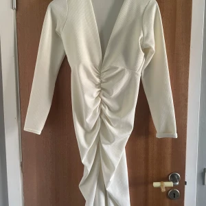 Vit långklänning med drapering - Snygg vit långklänning med djup v-ringning och långa ärmar. Klänningen har en cool draperad effekt framtill som ger en unik siluett. Tillverkad i ett strukturerat tyg som känns både modernt och stilrent. Perfekt för dig som vill sticka ut.