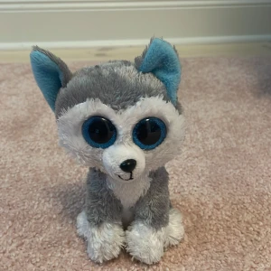 Ty Beanie Boo Slush - Ty Beanie Boo Slush är ett mjukt och charmigt gosedjur i form av en grå och vit hund med blå detaljer. Produkten ser ut att vara i gott skick utan synliga defekter eller slitage. Perfekt för samlare eller som present till barn.