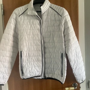 Vit quiltad pufferjacka med dragkedja - Snygg vit pufferjacka med svarta detaljer och horisontella quiltade sömmar. Jackan har hög krage, två fickor med dragkedja och svart piping längs axlarna. Perfekt för kyliga dagar och ger en clean look.