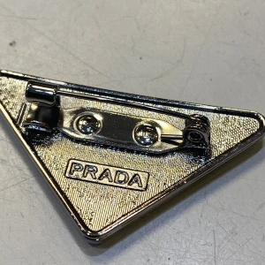 Triangelformad brosch  - Snygg triangelformad brosch i silverfärgad metall med vit emalj och tydlig logotyp. Broschen har klassisk nål på baksidan och detaljer med texten . Perfekt accessoar för att lyfta din outfit.