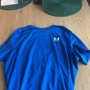 Blå Under Armour HeatGear t-shirt - Snygg blå t-shirt från Under Armour med HeatGear-teknologi. T-shirten har korta ärmar, rund hals och logga både fram och bak. Materialet är lätt och andas, perfekt för träning eller sportiga aktiviteter. Passformen är slim och ger en modern look.