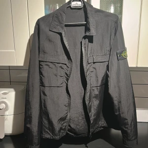 Svart overshirt från Stone Island - Svart overshirt från Stone Island med klassisk krage, två stora bröstfickor och dragkedja framtill. Ikonisk Stone Island patch på vänster ärm. Långärmad och tillverkad i lätt polyester som ger en clean och modern look.
