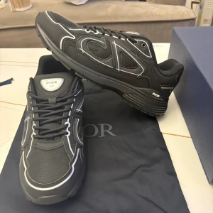 Svarta Dior B30 sneakers - Säljer ett par svarta Dior B30 sneakers med vita detaljer och stora CD-loggor på sidorna. Skorna har mesh och syntetmaterial, rund tå och snörning. Snygg och modern design som sticker ut, perfekt för dig som gillar exklusiva sneakers.