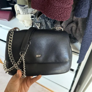 Svart axelväska från DKNY i skinn - Säljer en stilren svart axelväska från DKNY i mjukt skinn med silverfärgade kedjor som axelrem. Väskan har en enkel och klassisk design med DKNY-logga framtill. Perfekt storlek för det viktigaste och ger en snygg touch till din outfit.