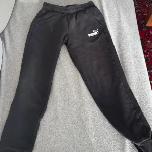 Svarta mjukisbyxor från Puma - Svarta mjukisbyxor från Puma med resår i midjan och muddar vid bensluten. Byxorna har två sidofickor och en vit Puma-logga på vänster lår. Perfekta för chill eller träning, tillverkade i mjukt material för extra komfort.