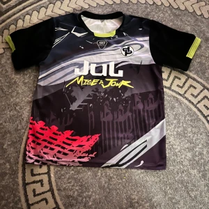 Svart och neon fotbollströja JUL Nineta - Snygg fotbollströja från JUL Nineta med coolt grafiskt tryck i svart, grått, vitt och neon-gult. Framsidan har texten 'Mise à Jour' och logga, baksidan har stor rund logga och texten 'Nineta'. Tillverkad i lätt och ventilerande polyester, perfekt för match eller träning.