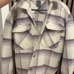 Rutig overshirt från Primark i lila och vitt - Säljer en oversized rutig overshirt från Primark i storlek S. Jackan är vit med lila rutor, har stora bröstfickor med lock och knäppning framtill. Perfekt för lager-på-lager och har en mjuk, ullig känsla. Snygg att slänga över en hoodie eller t-shirt.