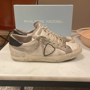 Philippe Model vita sneakers i skinn - Snygga vita sneakers från Philippe Model Paris med grå och svarta detaljer. Skorna är i skinn med mockadetaljer och har klassisk snörning samt rund tå. Bakpartiet är svart med logga och sidorna har den ikoniska skölden. Perfekta för en trendig streetstyle-look. Om de är något ni undrar hör av er direkt.