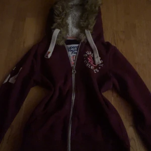 Vinröd Superdry hoodiejacka med fuskpäls - Säljer en vinröd hoodiejacka från Superdry med fluffig fuskpäls i luvan och vita snören. Jackan har dragkedja, tryck på bröstet och siffror på ärmen. Perfekt för dig som gillar lite y2k stil den passar mig jätte bra och jag har xs/s 