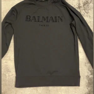 Svart sweatshirt från Balmain Paris - Snygg svart sweatshirt från Balmain Paris med broderad logga framtill. Tröjan har en klassisk rund halsringning och långa ärmar. Perfekt för dig som gillar stilrena designer och exklusiva märken. Passar 15-17 åringar som väger cirka 70 Kg.