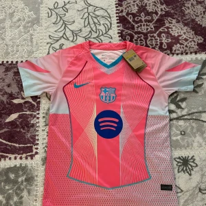 Barça x Nike rosa fotbollströja  - FC Barcelona Dri-Fit fotbollströja från Nike i rosa och ljusblått med blå detaljer. Tröjan har korta ärmar, V-ringning med blå kant, klubbmärke och Spotify-logga på bröstet. Tillverkad i lätt och ventilerande polyester. Fraktar inom 24 timmar.