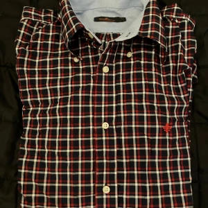 Rutig skjorta från Morris Stockholm - Snygg rutig skjorta från Morris Stockholm i mörkblått, rött och vitt. Klassisk krage, vita knappar och långärmad modell. Broderad röd logga på bröstet. Skjortan är i bomull och har en normal passform, perfekt för dig som gillar stilrena detaljer.