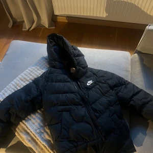 Svart pufferjacka från Nike med huva - Säljer en svart pufferjacka från Nike med huva och dragkedja. Jackan har en clean look med diskret Nike-logga på bröstet och är perfekt för kyliga dagar. Passar dig som gillar sportig och enkel stil.