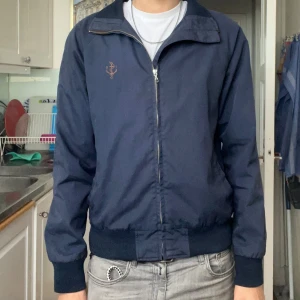 Mörkblå vindjacka med dragkedja - Snygg mörkblå vindjacka med dragkedja framtill och ribbade muddar vid ärmslut och midja. Jackan har en liten broderad ankare-detalj på bröstet och klassisk krage. Perfekt för dig som gillar stilren och enkel design.