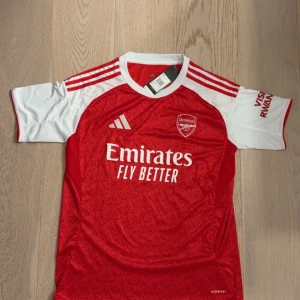 Arsenal hemmatröja 25/26 - Arsenal hemmatröja 25/26 från Adidas i rött och vitt med klubbmärke och sponsortryck på bröstet. Tröjan har korta ärmar, vita axelpartier och röda Adidas-ränder. Tröjan är helt ny med snygga små ”A” detaljer på hela tröjan