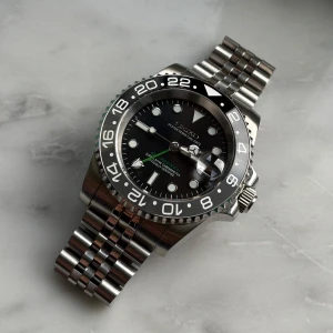 Seiko “Bruce Wayne” GMT-mod - Seiko “Bruce Wayne” GMT-mod! Klockan har en 40 mm boett, automatisk NH34-verk med fullt fungerande GMT-funktion. Utrustas med safirglas, keramisk bezel samt genomskinlig bakboett. Skruvad krona, 3 ATM vattentålighet, stark Luminova och justerbar länk (14,5–22 cm).  Skicka DM för fler bilder och info!