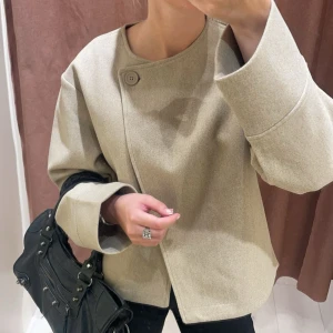 Beige omlott kappa med knapp - Stilren beige kappa med omlottknäppning och en synlig knapp vid halsen. Jackan har en boxig, oversized passform och är tillverkad i ett mjukt ullmaterial. Den har rund halsringning och långa ärmar, perfekt för höst och vinter.