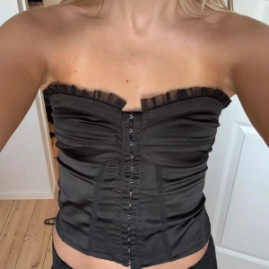 Svart korsett från Gina Tricot - Supersnygg svart korsett från Gina Tricot med volangdetalj upptill och hyskknäppning framtill. Gjord i en blandning av polyester och bomull med lite elastan för stretch. Axelbandslös och figurnära passform, perfekt att styla till olika looks.