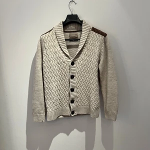 Beige kofta från LC WAIKAKIKI M - Säljer en beige stickad kofta från LC WAIKAKIKI i storlek M. Koftan har grovt stickat mönster framtill, bruna mockadetaljer på axlar och armbågar samt bred ribbad krage. Stängs med bruna knappar och har långa ärmar. Perfekt för lager-på-lager-stil. Hör av er vid frågor 