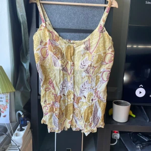 Blommig gul linnetopp med spetsdetaljer - Säljer en söt linnetopp med blommigt mönster i gult, vitt och lila. Toppen har smala axelband, spetsdetaljer vid urringningen och en lätt rynkad nederkant. Materialet är mjukt och luftigt, perfekt för varma dagar. Mönstret består av blad och blommor i olika nyanser.