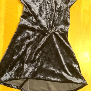 Svart sammetsklänning med knut - Mörk sammetsklänning i svart med kort längd och snygg knutdetalj framtill. Klänningen har en mjuk, glansig yta som ger en lyxig känsla och passar perfekt när du vill sticka ut. Enkel men ändå elegant design.