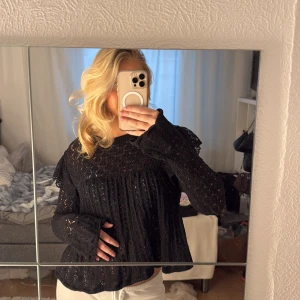 Svart spetsblus med volang - Säljer en svart blus med vackra spetsdetaljer och volang vid axlarna. Blusen har långärmade, lätt utsvängda ärmar och ett luftigt, romantiskt fall. Perfekt för dig som gillar en feminin och trendig look. Storlek XL men passar mig som brukar ha S/M