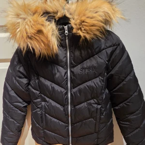 Svart pufferjacka från Svea - Svart pufferjacka från Svea med snyggt broderad logga på bröstet och patch på ärmen. Jackan har en stor huva med fluffig fuskpäls och dragkedja framtill. Snyggt chevronmönster i vadderingen och lång ärm. Som ny!