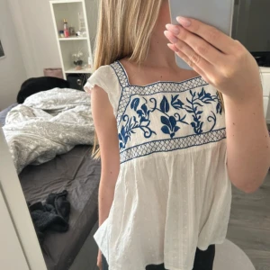 Vit blus med blå broderi och volang - Supersöt vit blus med blå broderade blommor upptill och volangdetaljer vid axlarna. Blusen har knytning i ryggen och är ärmlös med en luftig, flowy passform. Perfekt för varma dagar och ger en romantisk vibe. nästan aldrig  använd