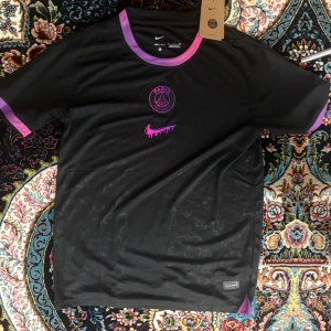 Svart PSG fotbollströja Nike - Snygg svart PSG fotbollströja från Nike med lila och rosa detaljer vid krage och ärmslut. Tröjan har klubbmärke och Nike-logga i rosa framtill. Tillverkad i lätt och ventilerande material, perfekt för träning eller match.