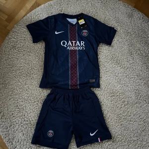 PSG fotbollströja och shorts Nike - Säljer ett PSG matchställ från Nike med Dri-Fit teknologi. Tröjan är marinblå med korta ärmar, har ett randigt mönster i rött och vitt längs mitten, PSG-logga och Qatar Airways tryck. Materialet är lätt och andas, perfekt för träning eller match.