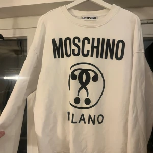Vit sweatshirt från Moschino - Säljer en vit sweatshirt från Moschino med stor svart logga och tryck framtill. Tröjan har rund halsringning och långa ärmar, perfekt för dig som gillar streetstyle och vill sticka ut med ett ikoniskt märke.