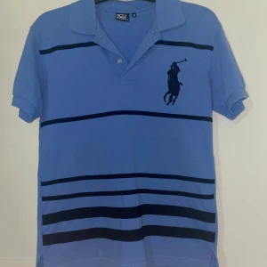 Blå randig piké från Polo Ralph Lauren - Snygg blå pikétröja från Polo Ralph Lauren med mörkblå ränder och klassisk polospelare broderad på bröstet. Tröjan har korta ärmar, krage och knappar vid halsen. Perfekt för en chill och stilren look.