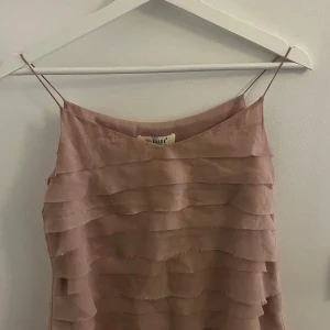 Ljusrosa volang linnetopp från Elise - Supersöt ljusrosa linnetopp från Elise i storlek S. Toppen har tunna axelband och flera lager med volanger som ger en cool och romantisk vibe. Perfekt för dig som gillar feminina detaljer och en luftig känsla. Materialet är mjukt och har en lätt glans.